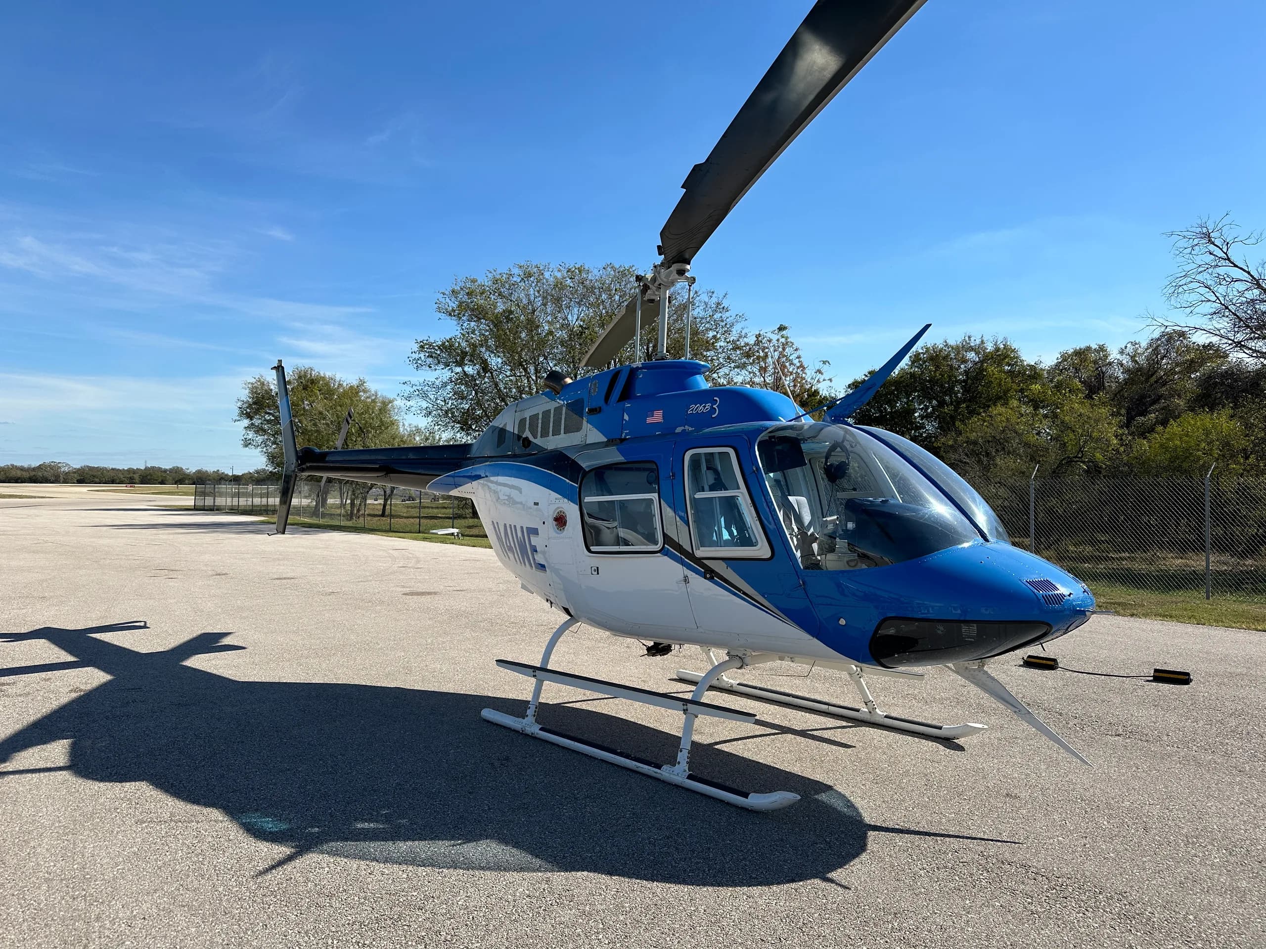 A blue and white Bell 206B3 helicopter with black accents is parked on an asphalt ramp under a clear blue sky.