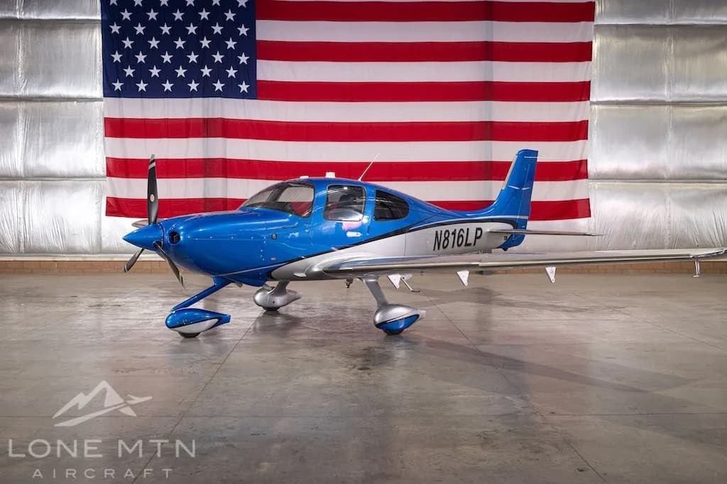 A blue and silver Cirrus SR22 aircraft, registration N816LP, parked in a hangar with an American flag in the background.