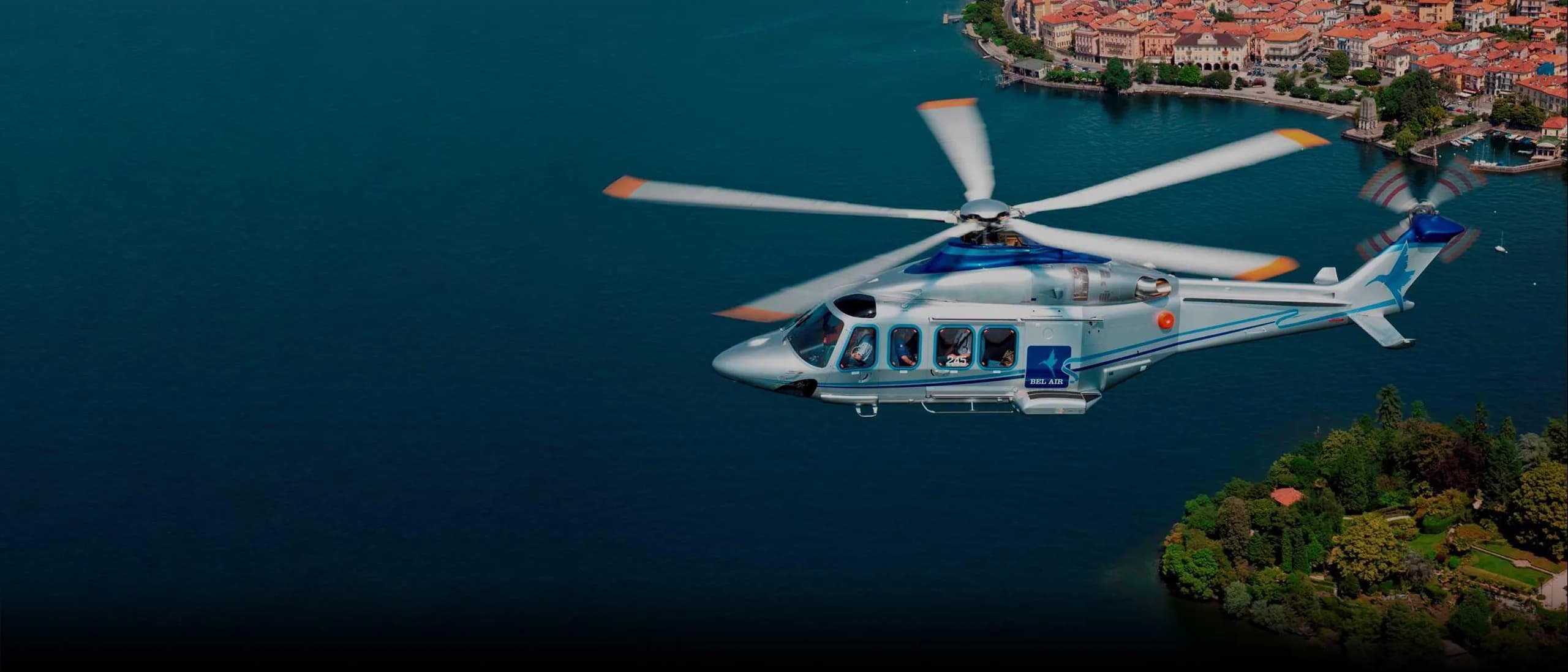 AgustaWestland AW169 helicopter flying over a lake near a town.