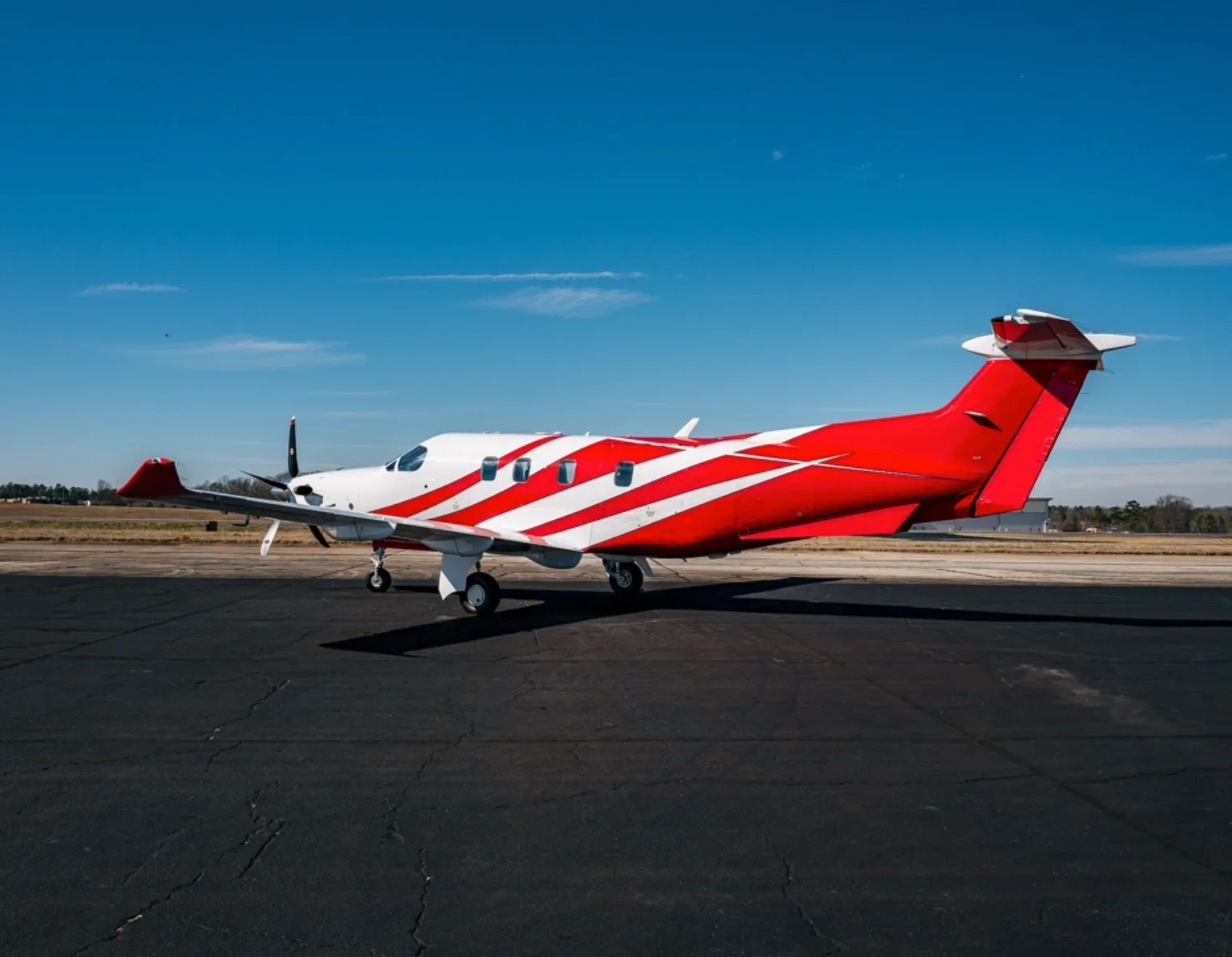 A Pilatus PC-12 turboprop aircraft with a red and white paint scheme parked on an airfield under a clear blue sky.