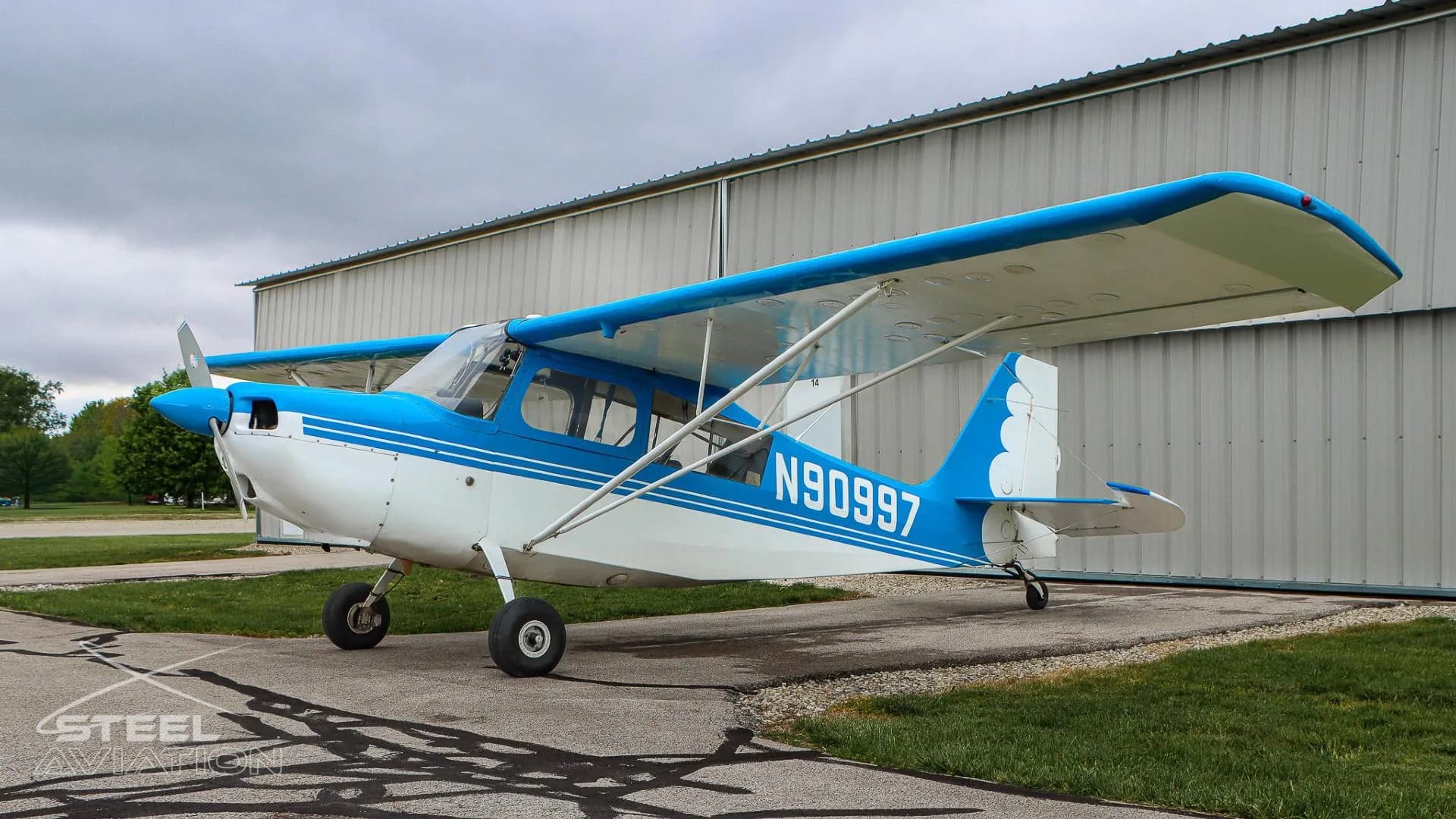 Side view of a white and blue Bellanca 7GCBC Citabria aircraft with registration N90997, parked in front of a hangar.