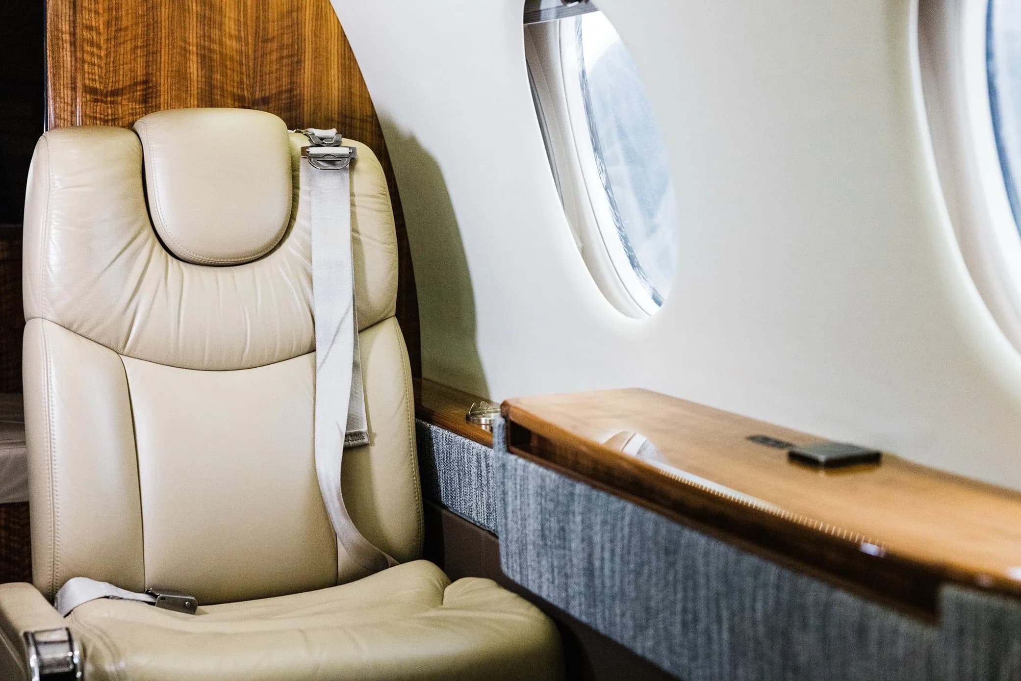 A beige leather seat next to a window in the private jet's cabin, with a wooden side table.