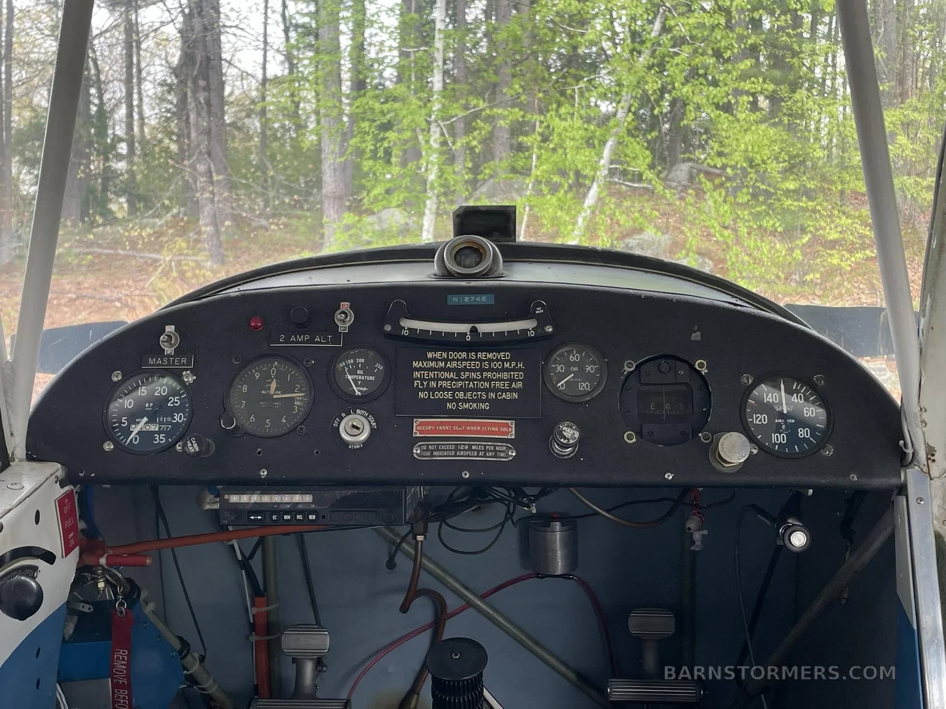 The cockpit instrument panel of a vintage light aircraft, showing various gauges and controls, with trees visible through the windshield.