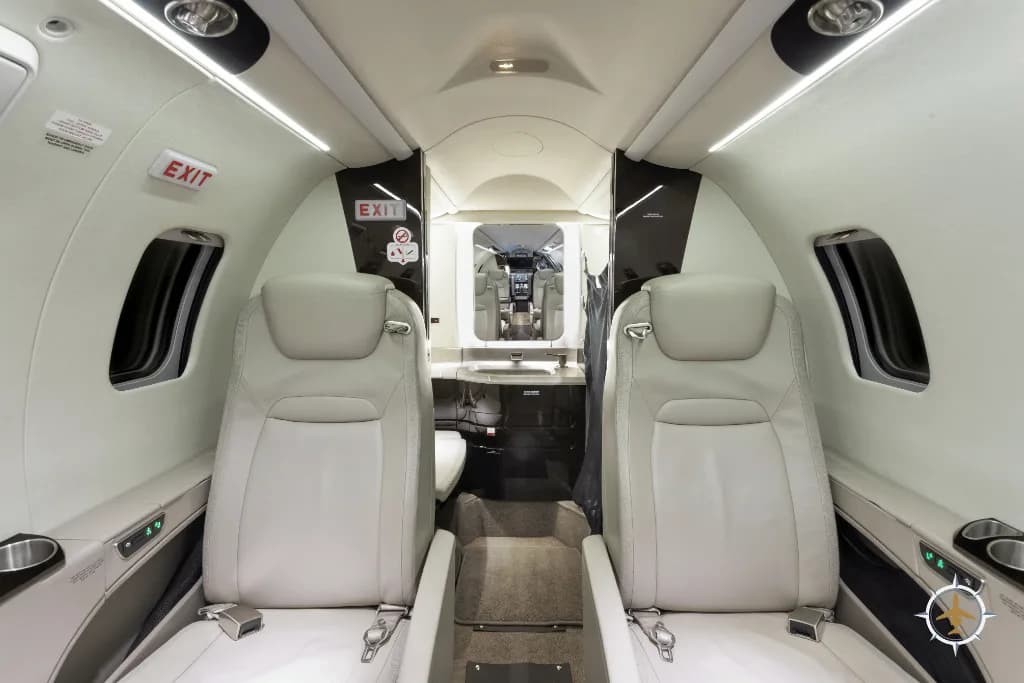 View of a luxurious aircraft cabin with two light-colored leather seats in the foreground and the cockpit visible in the distance.