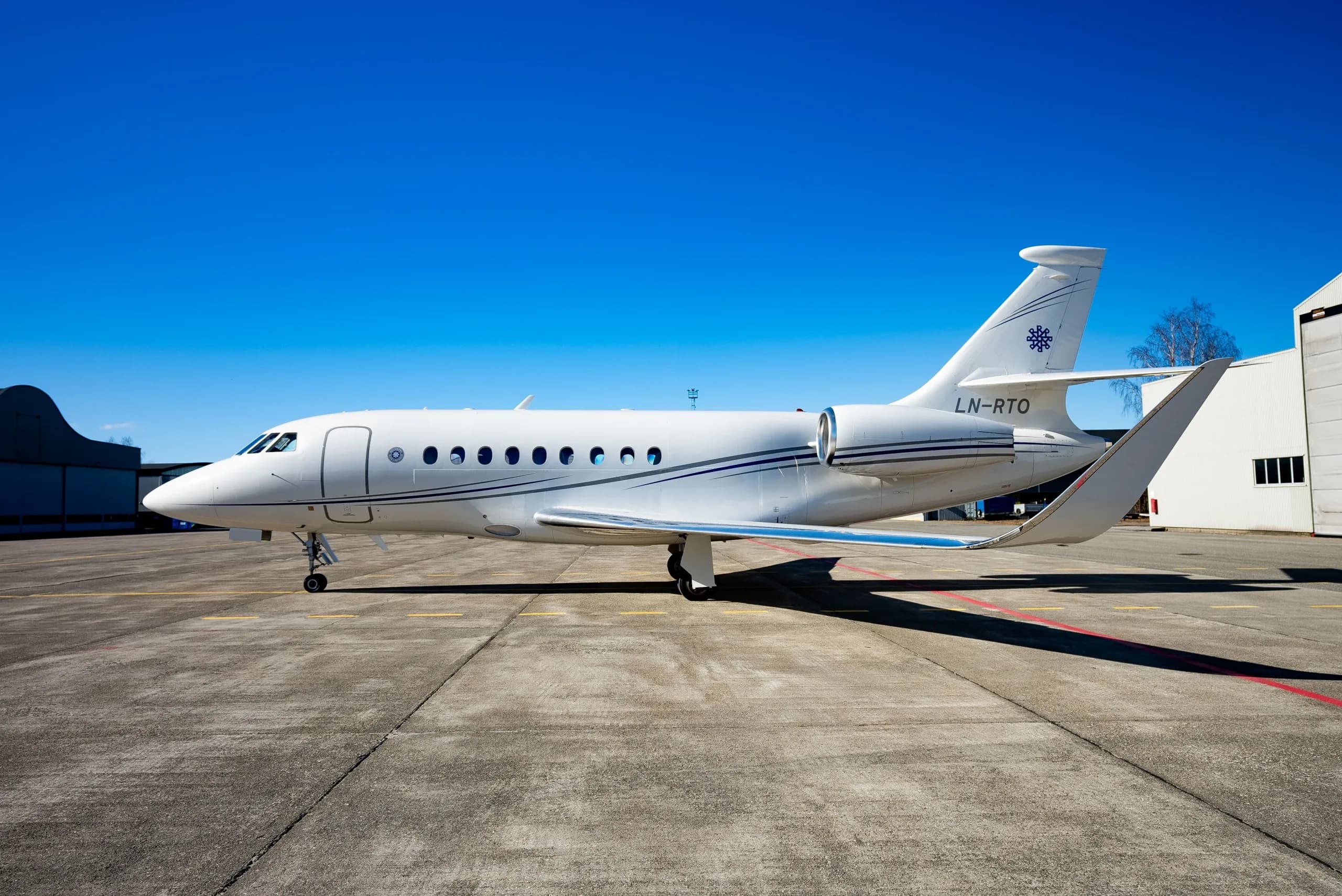 A white Dassault Falcon business jet with blue stripes and registration LN-RTO parked on a tarmac under a clear blue sky.