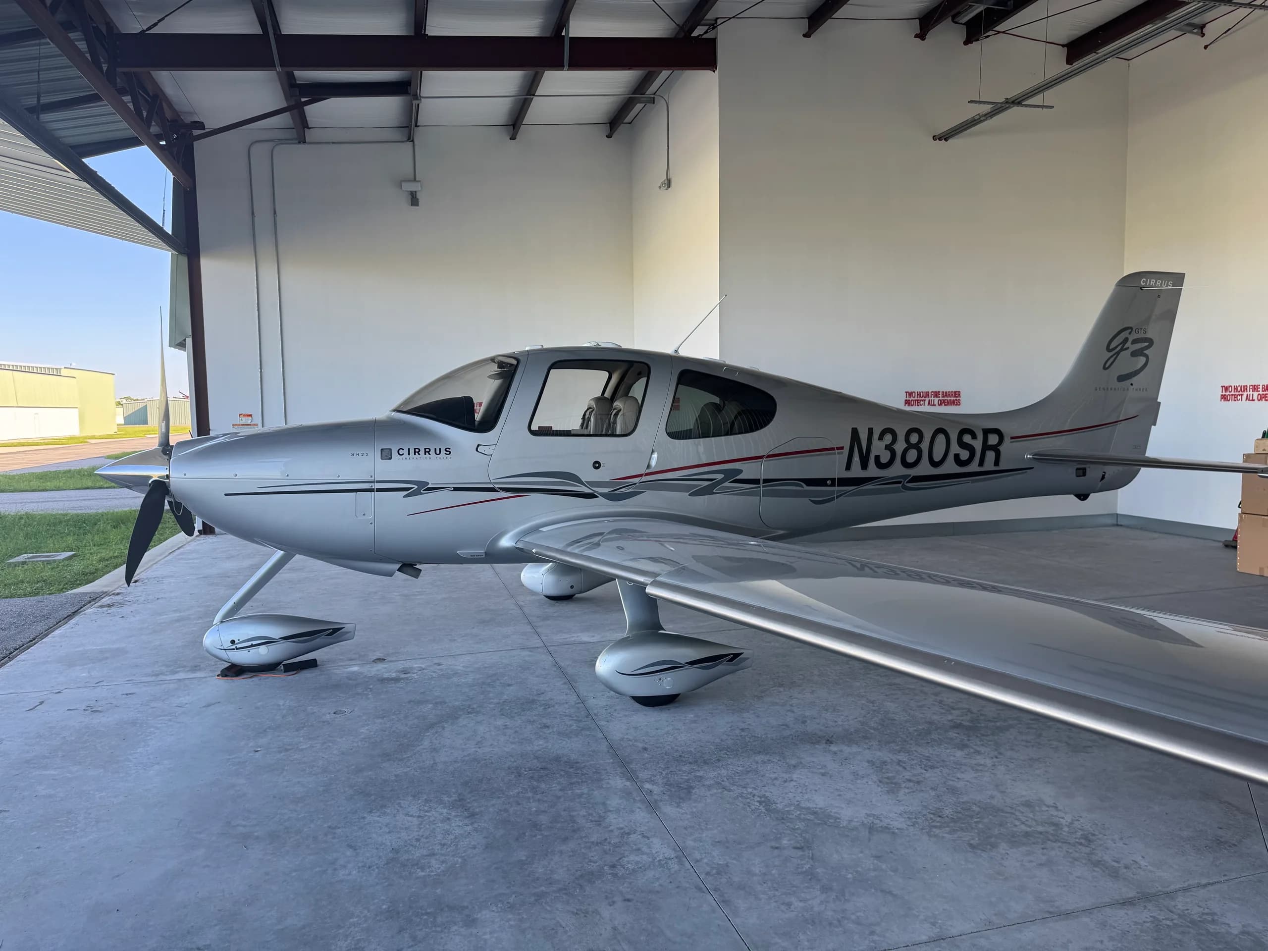 A silver Cirrus SR22 Generation Three aircraft with N380SR registration is parked in a hangar, viewed from the left side.