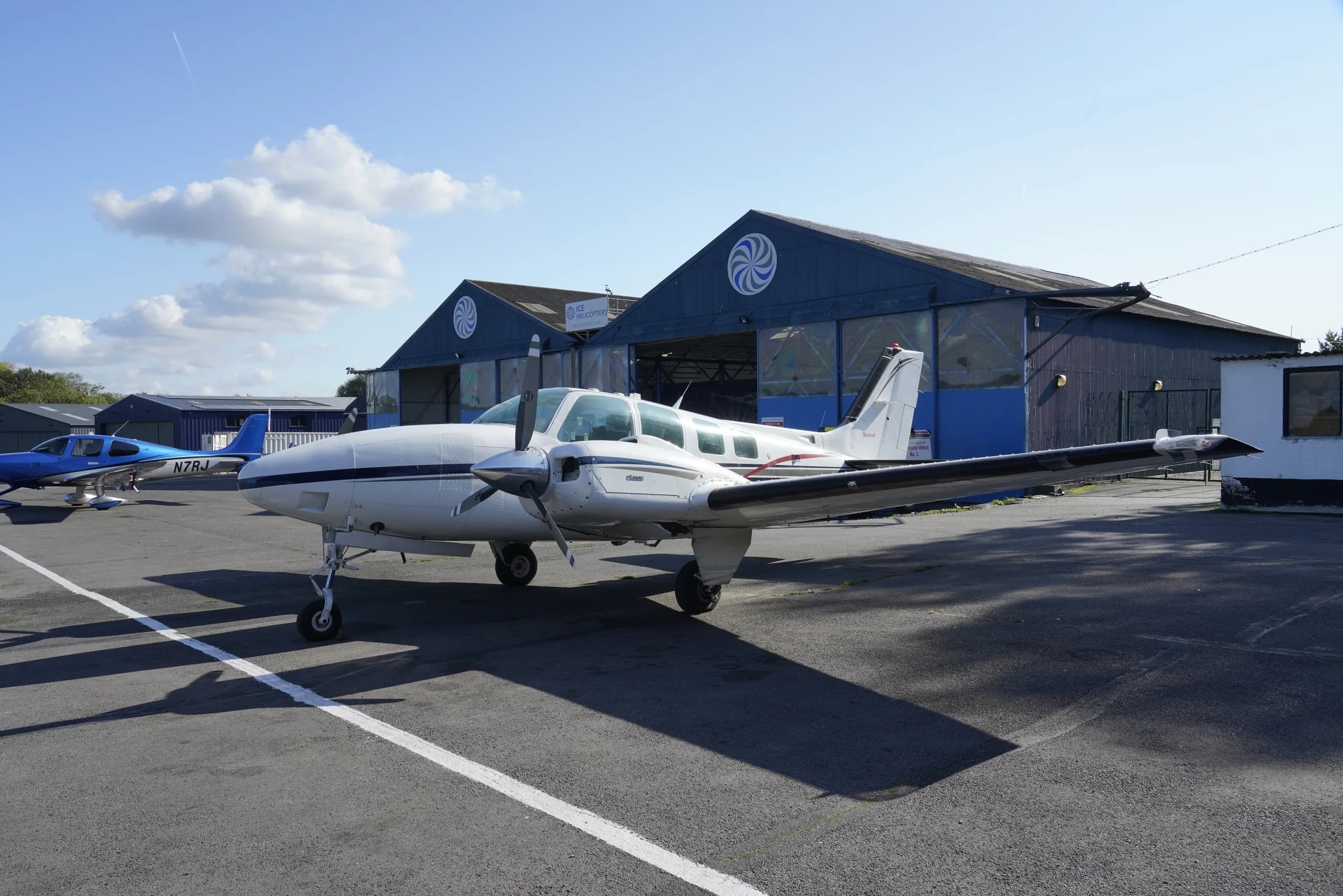 A white twin-engine propeller aircraft with blue and red stripes, registration N7223Y, parked on an asphalt apron with hangars and other aircraft in the background.