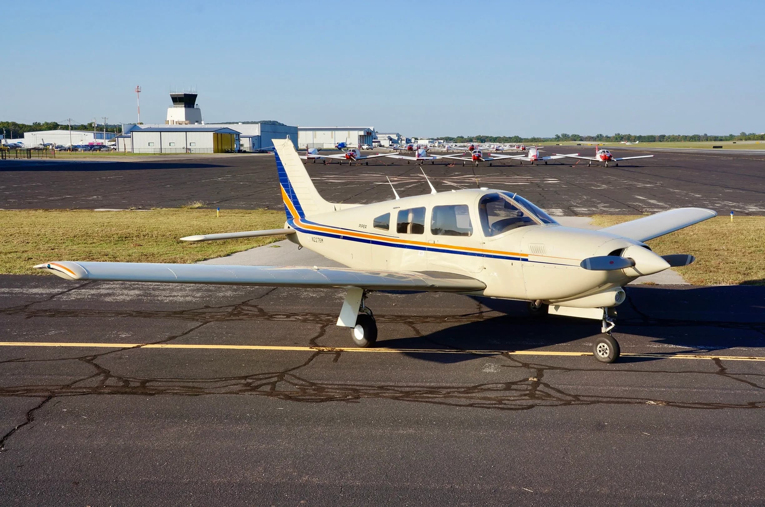 A cream-colored Piper aircraft with blue and orange stripes parked on an asphalt tarmac with a control tower in the background.