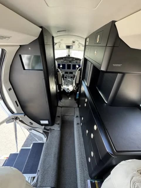A view down the aisle of a business jet, showing dark storage units on both sides leading to the cockpit.