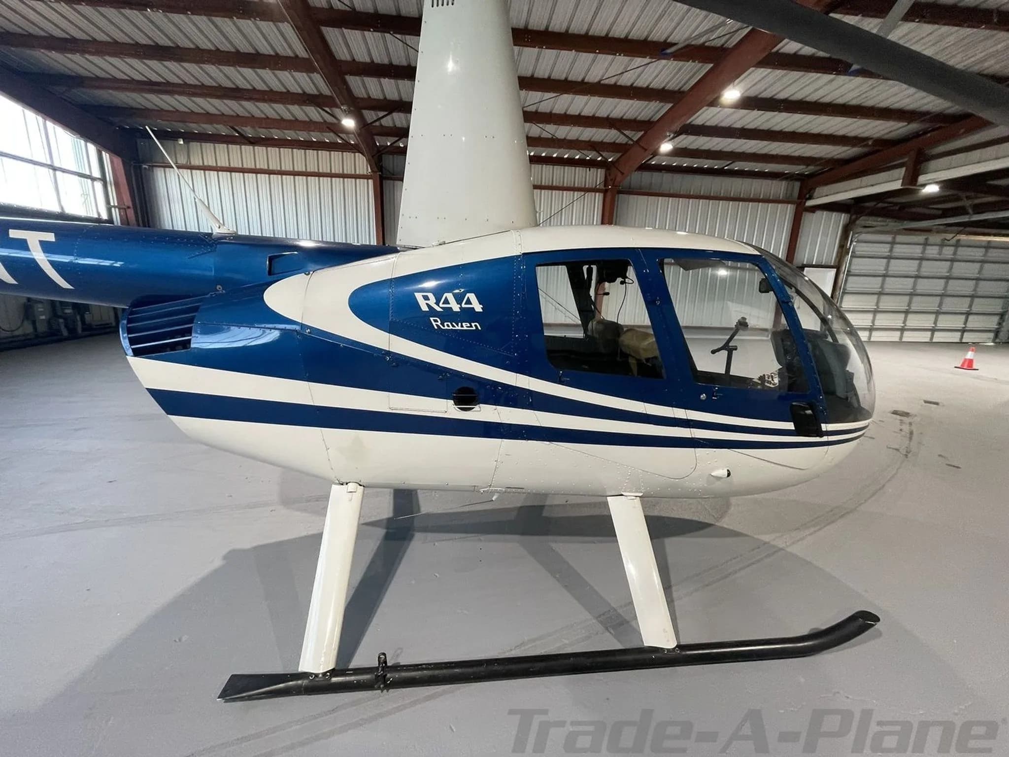 A close-up side view of the blue and white Robinson R44 Raven helicopter's fuselage in a hangar.