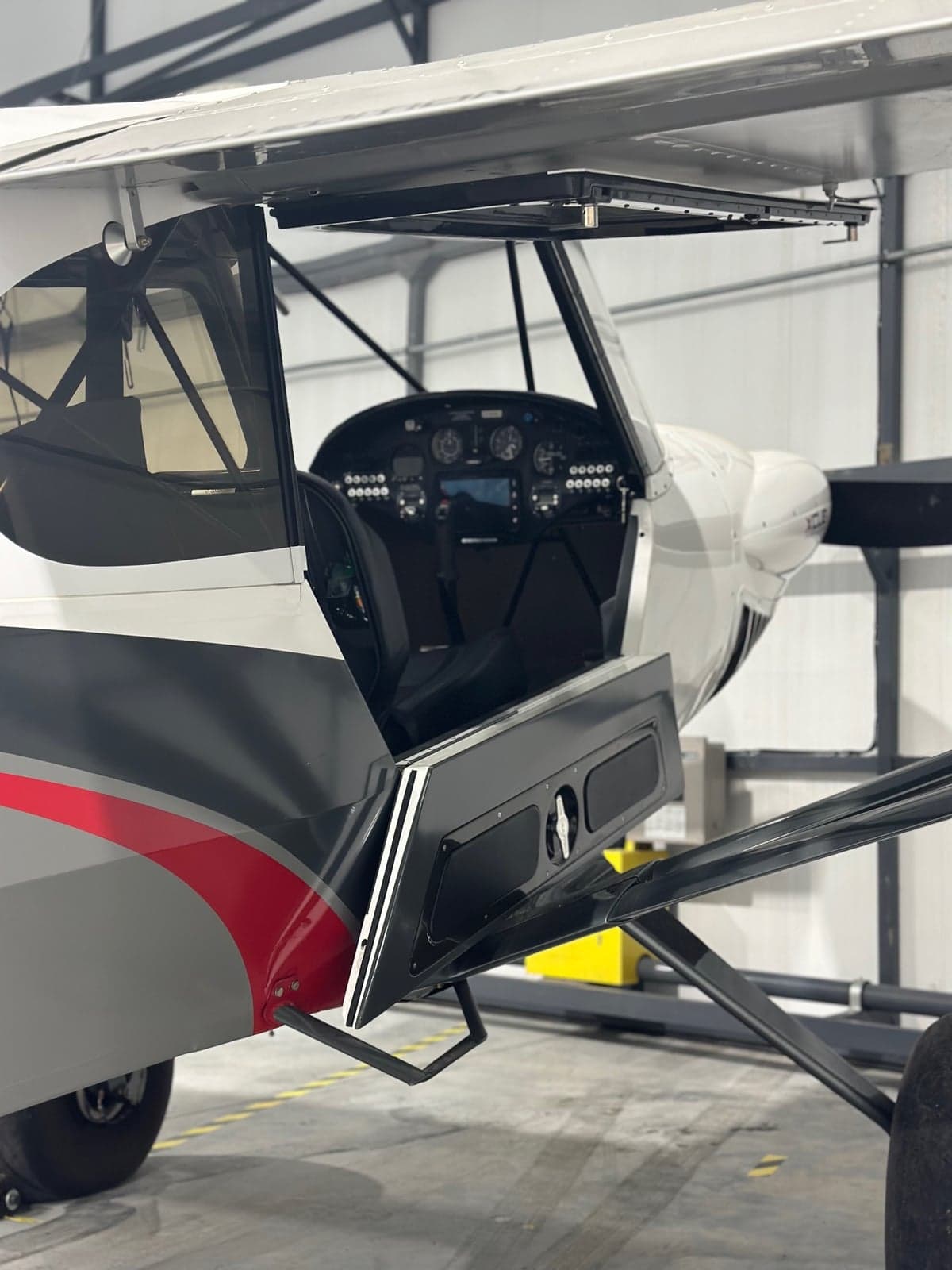 A white, grey, and red CubCrafters XCub aircraft with its door open in a hangar, revealing the cockpit.