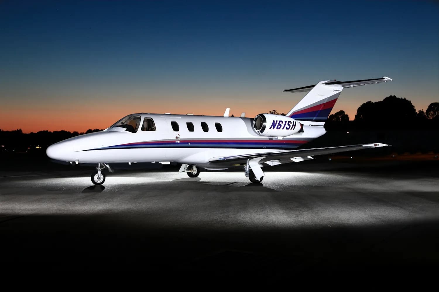 A white private jet with red, blue, and grey stripes on the fuselage and tail, illuminated on an airfield at dusk.