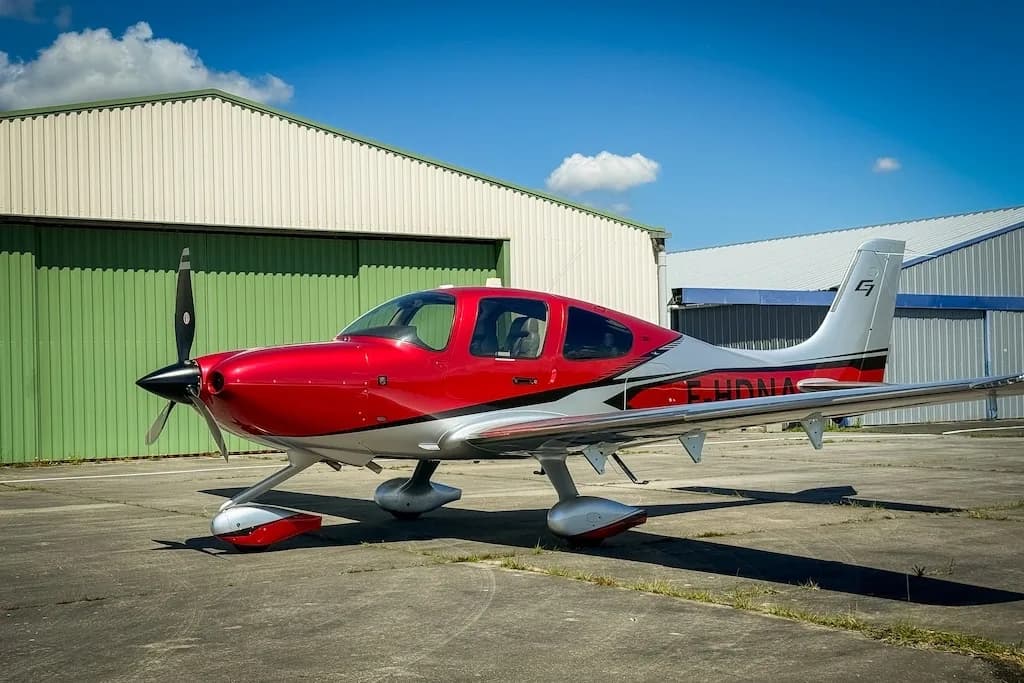 A red and silver Cirrus SR22 GTS (F-HDNA) aircraft parked on a tarmac in front of hangars under a blue sky.