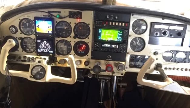 The cockpit instrument panel of a small aircraft, featuring analog gauges, digital displays, and two control yokes.