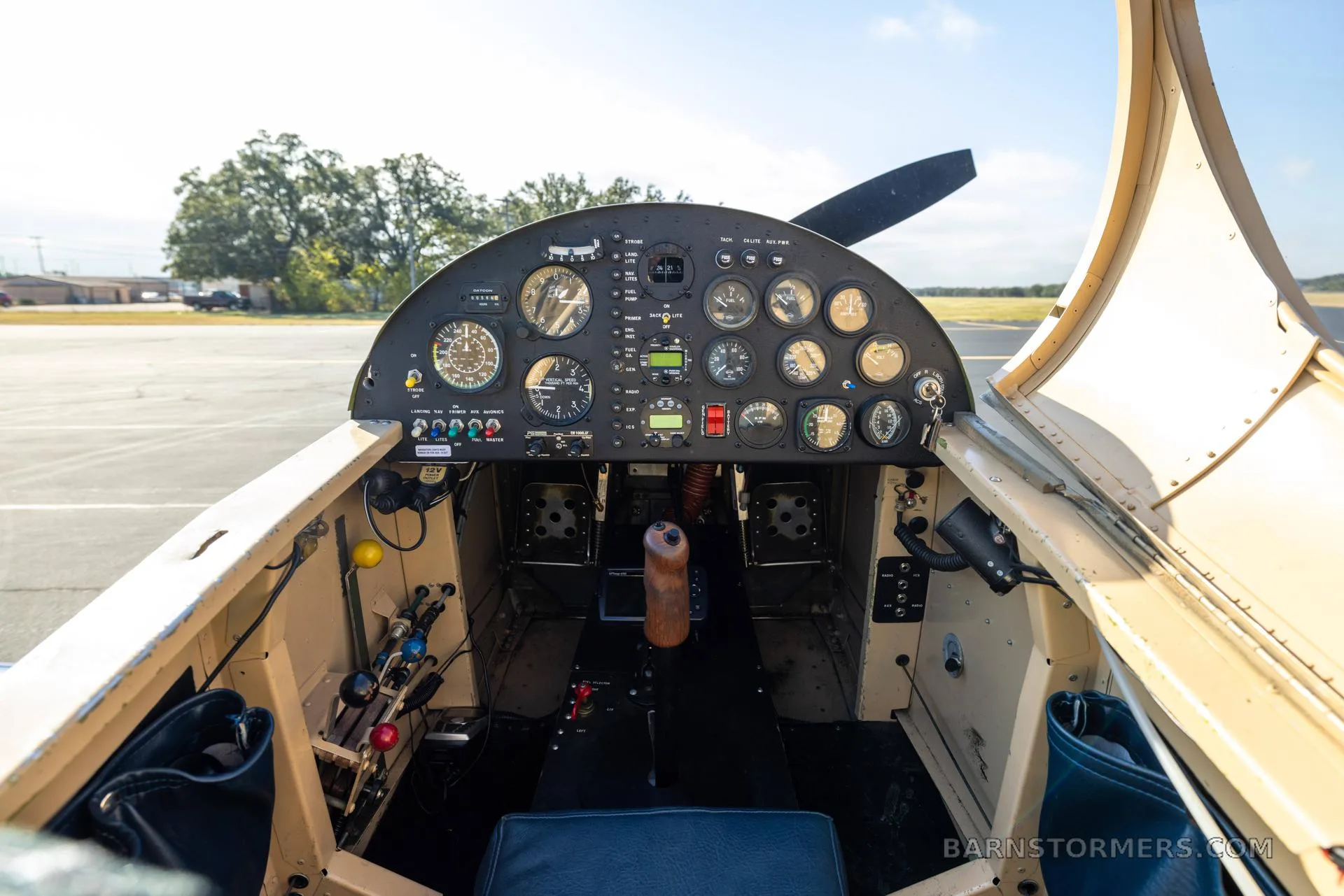 The cockpit of a single-seat aircraft showing the instrument panel, control stick, and blue and tan seat.