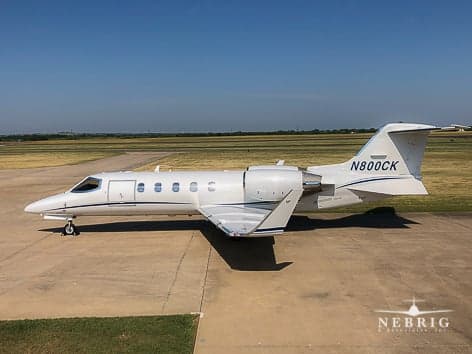 A white Learjet N800CK with blue stripes is parked on an airfield tarmac on a sunny day.