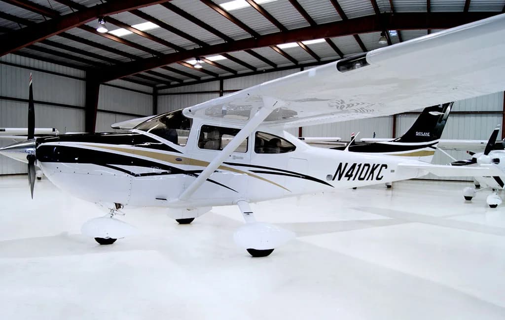 A white Cessna Skylane aircraft with black and gold stripes is parked in a hangar, viewed from the left side.