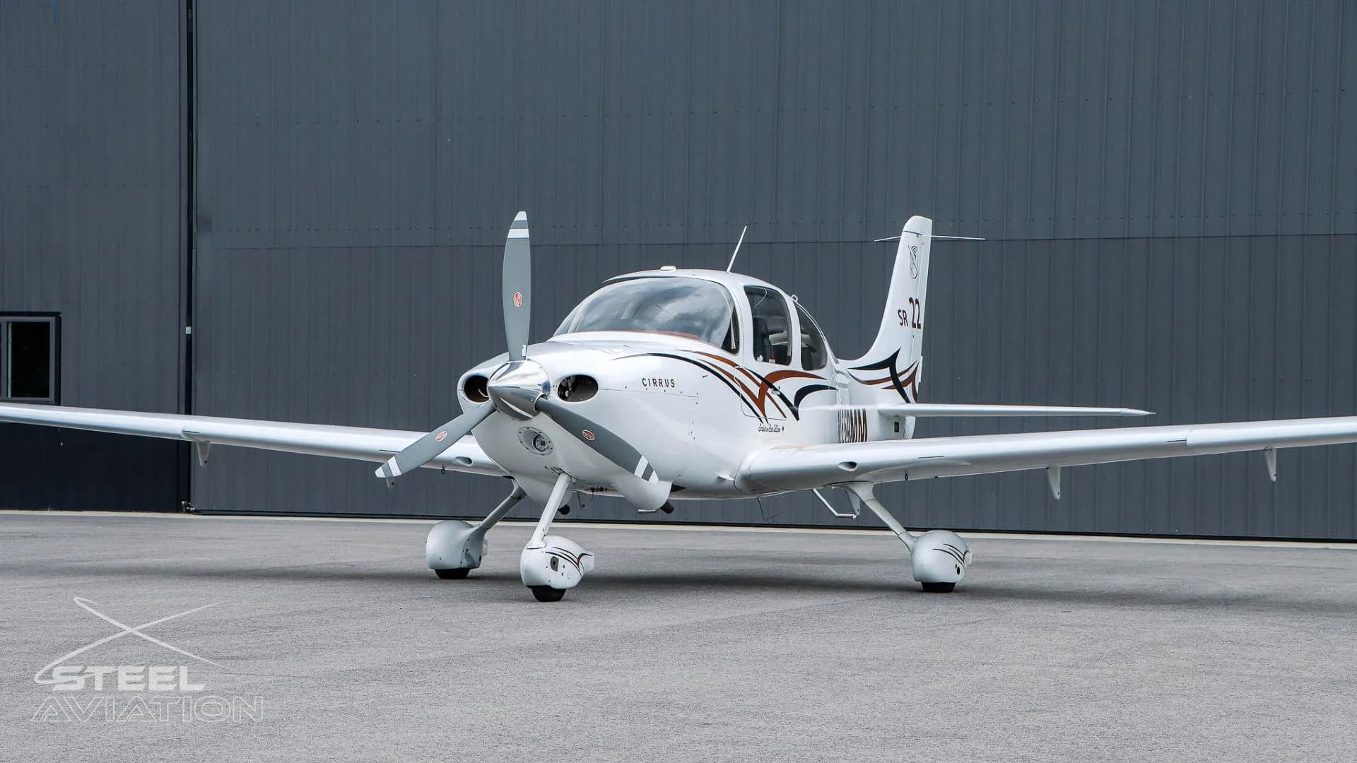 A white Cirrus SR22 aircraft with brown and black accent stripes is parked at an angle in front of a dark grey hangar.