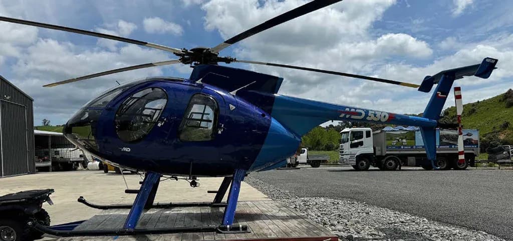 A blue and light blue MD 530 helicopter on a trailer, with a white truck and building in the background.
