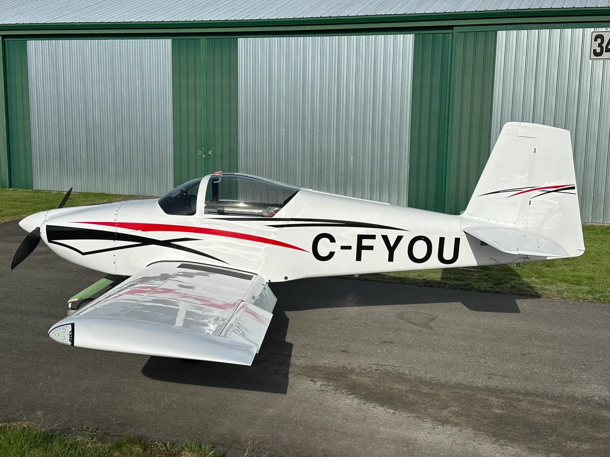 A white single-engine aircraft with red and black stripes on the fuselage and tail, registration C-FYOU, parked on an asphalt apron next to a green and silver hangar.