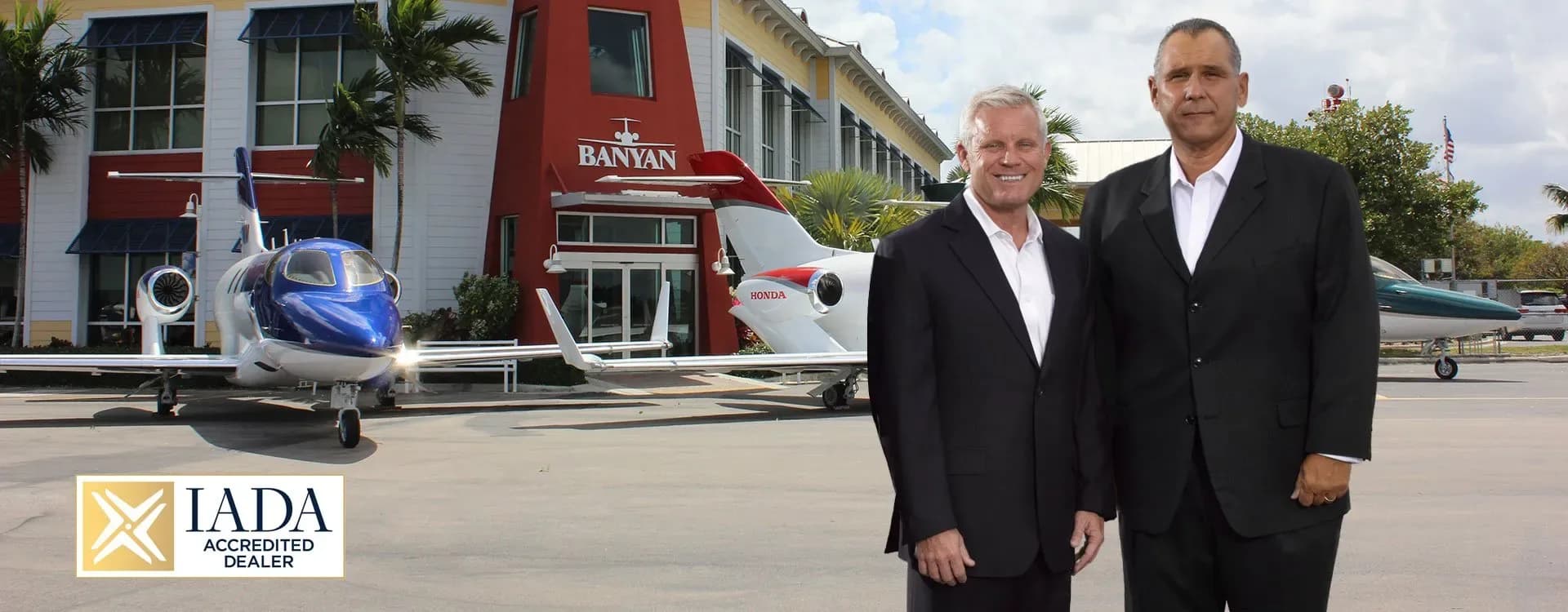 Two men stand in front of a Banyan building with two HondaJet aircraft on the tarmac.