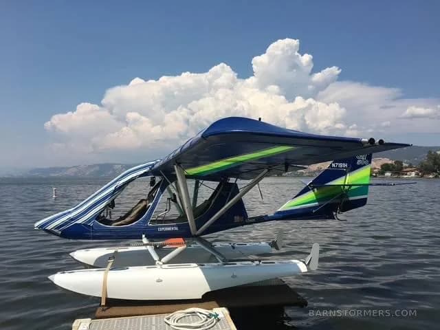 A blue and green experimental seaplane with white floats is docked on a calm lake under a partly cloudy sky.