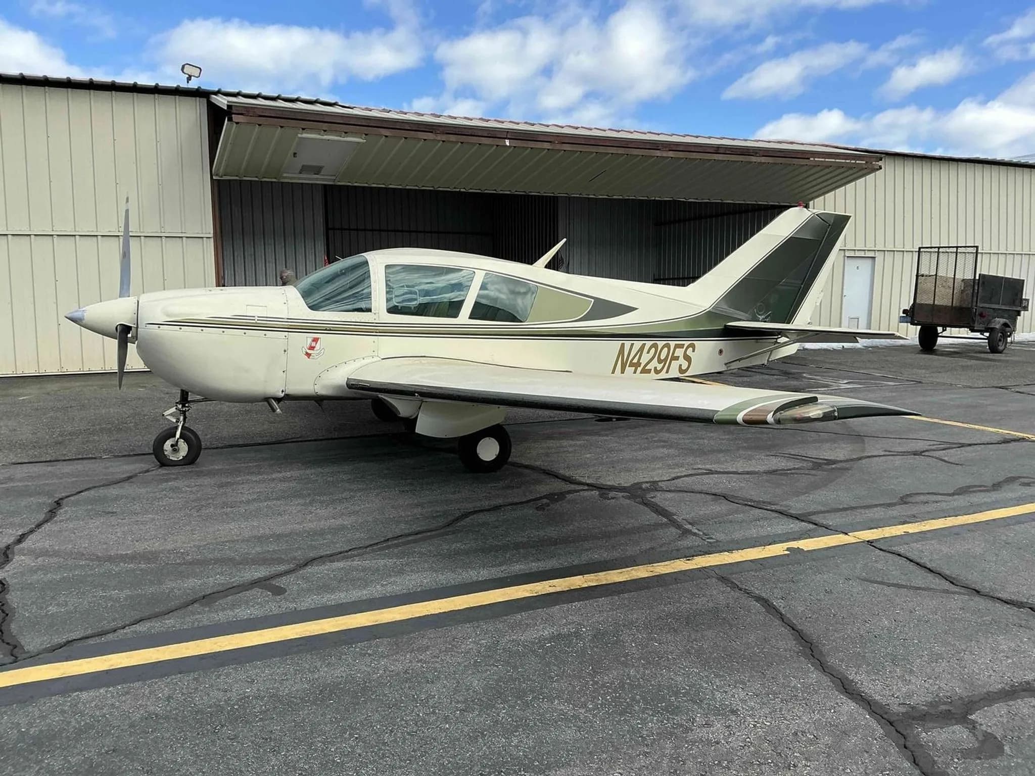 A side view of the white and green Bellanca Viking aircraft, N429FS, parked on an asphalt ramp near an open hangar.