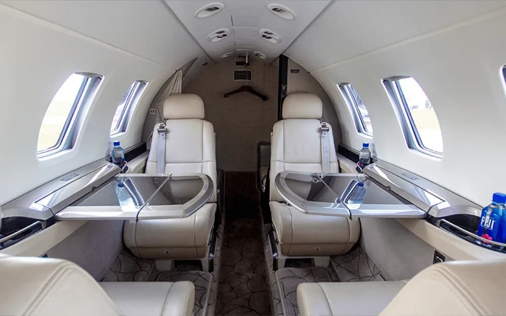 The aircraft cabin shows two rows of light-colored leather seats with fold-out tables and Fiji water bottles.