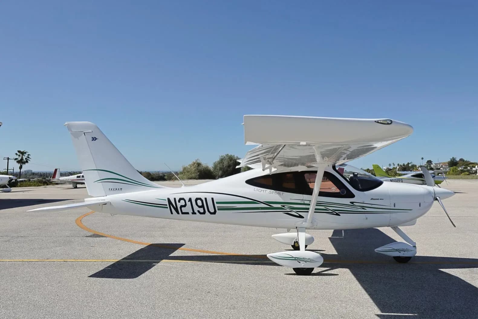 A white Tecnam P2008 Light Sport aircraft with green stripes, registration N219U, parked on an airport apron under a clear sky.