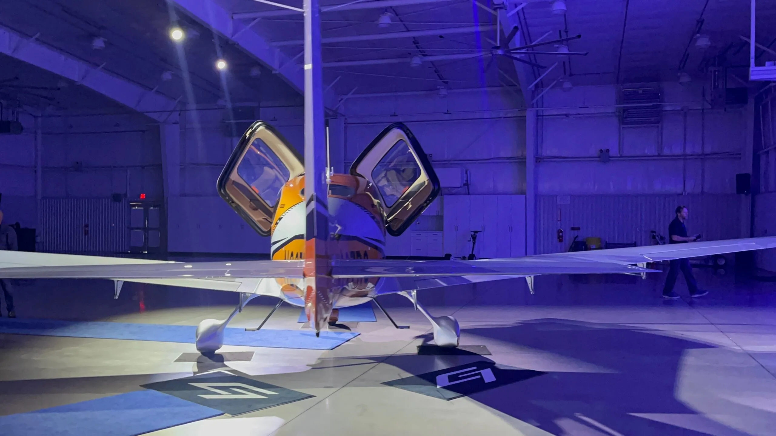 A front-rear view of a Cirrus SR22 aircraft with orange and silver paint, doors open, in a blue-lit hangar.