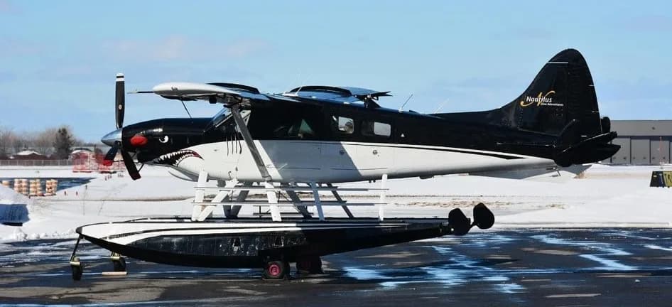 A black and white seaplane with a shark mouth paint scheme on the nose, parked on a snowy tarmac.