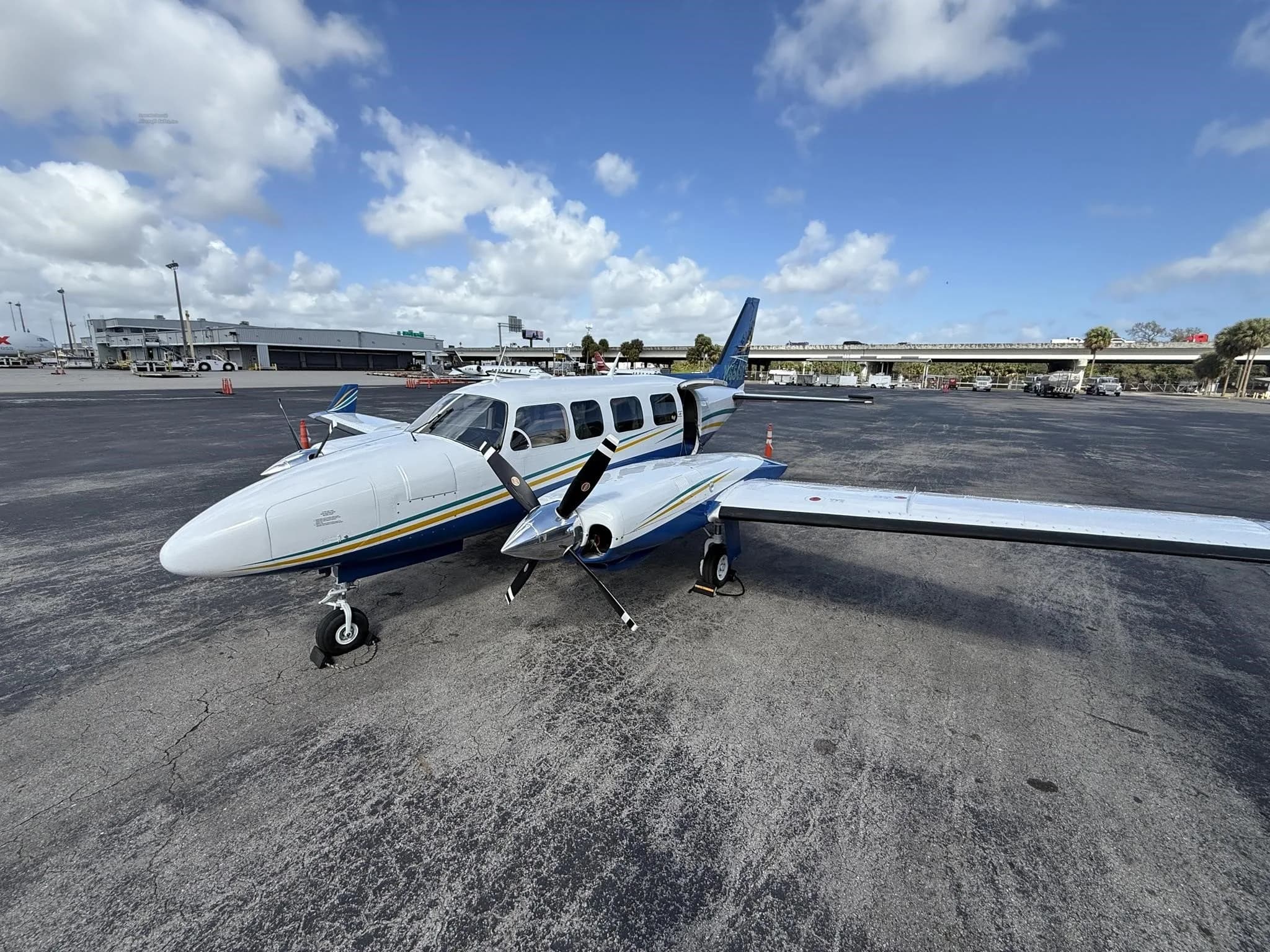 A white and blue twin-engine propeller aircraft with yellow and green stripes, parked on an airport tarmac with its passenger door open.