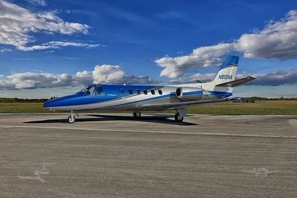 A blue and white private jet, tail number N812HA, is parked on an asphalt runway under a partly cloudy sky.