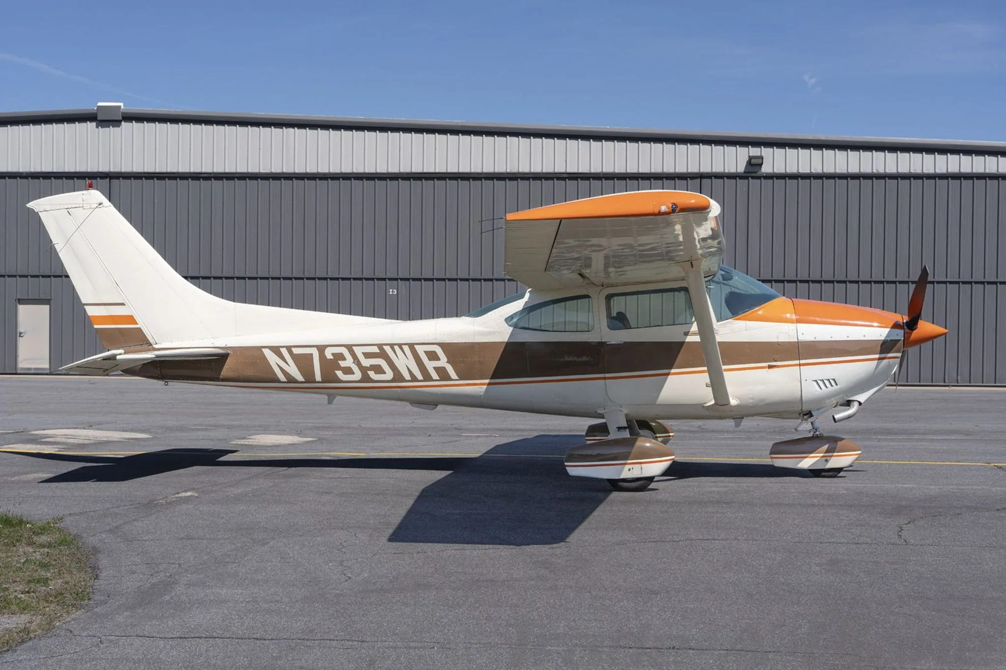 A full side profile of a white and brown Cessna 172 with orange accents, parked on an asphalt ramp.