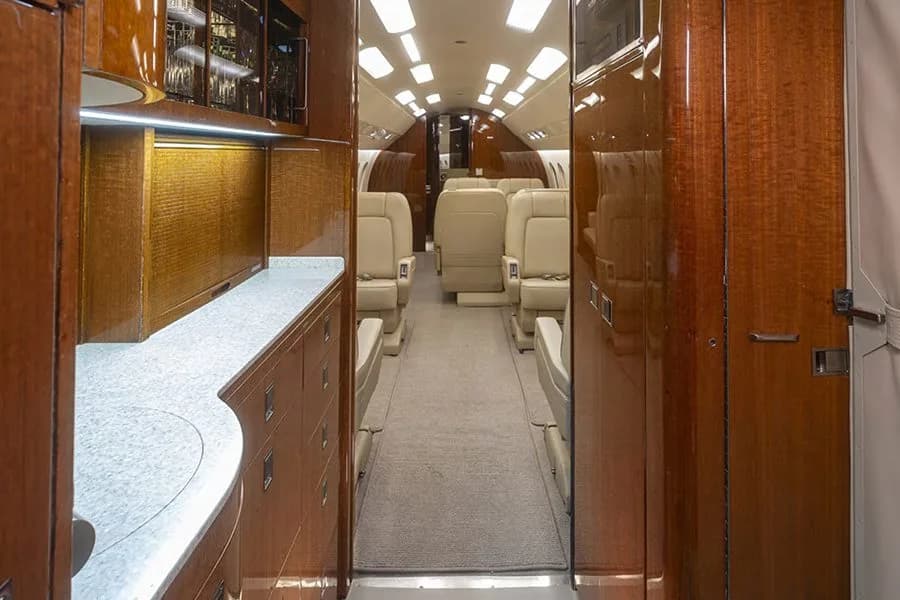 The galley area of a private jet, featuring dark wood cabinetry, a light-colored countertop, and a view down the cabin aisle.