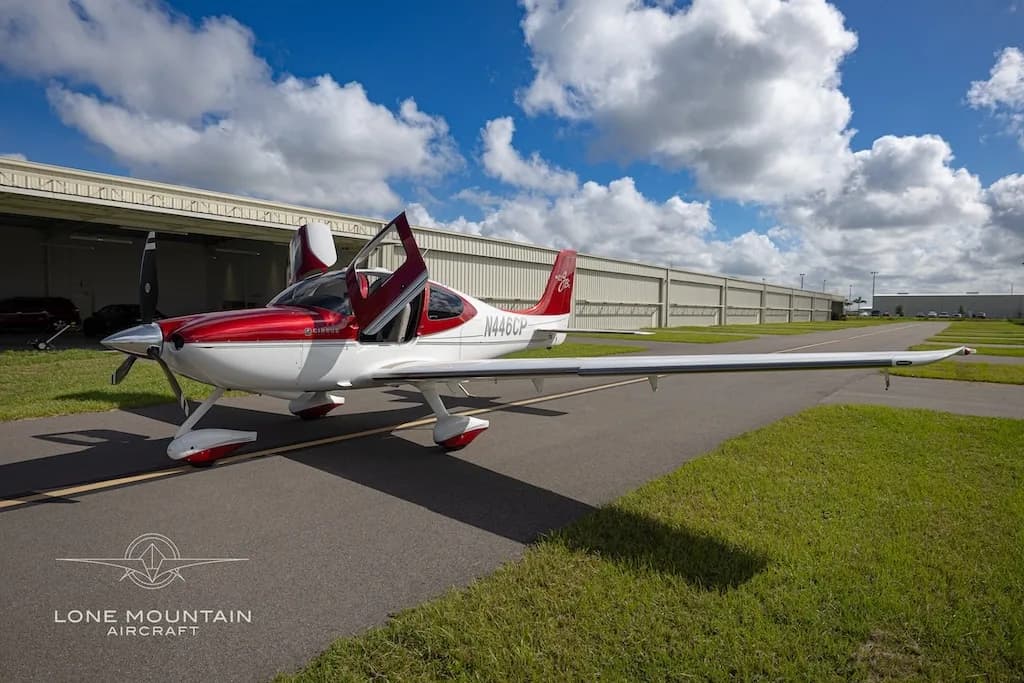 A red and white Cirrus SR22 aircraft (N446CP) with its doors open, parked on an asphalt ramp with hangars in the background.