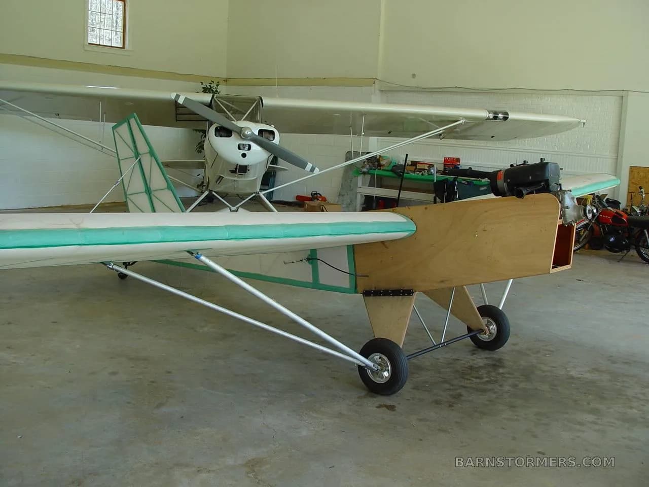 A three-quarter front view of a light aircraft with a wooden fuselage and green trim, parked in a hangar.