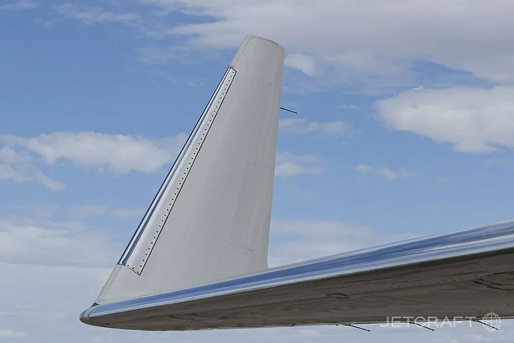 A close-up view of the wingtip of a white Bombardier Global 6000 business jet, showing blue and grey stripes against a blue sky.