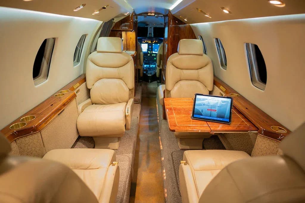 The aircraft's interior cabin with beige leather seats, wood trim, and a tablet on a fold-out table.
