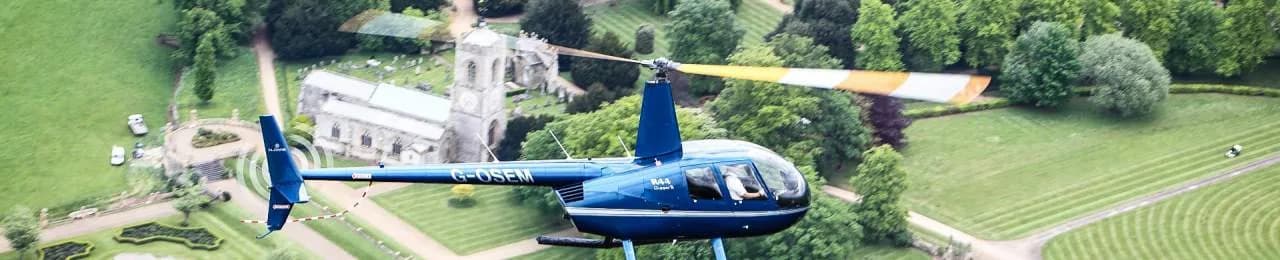 A blue Robinson R44 helicopter flying over a church and gardens.