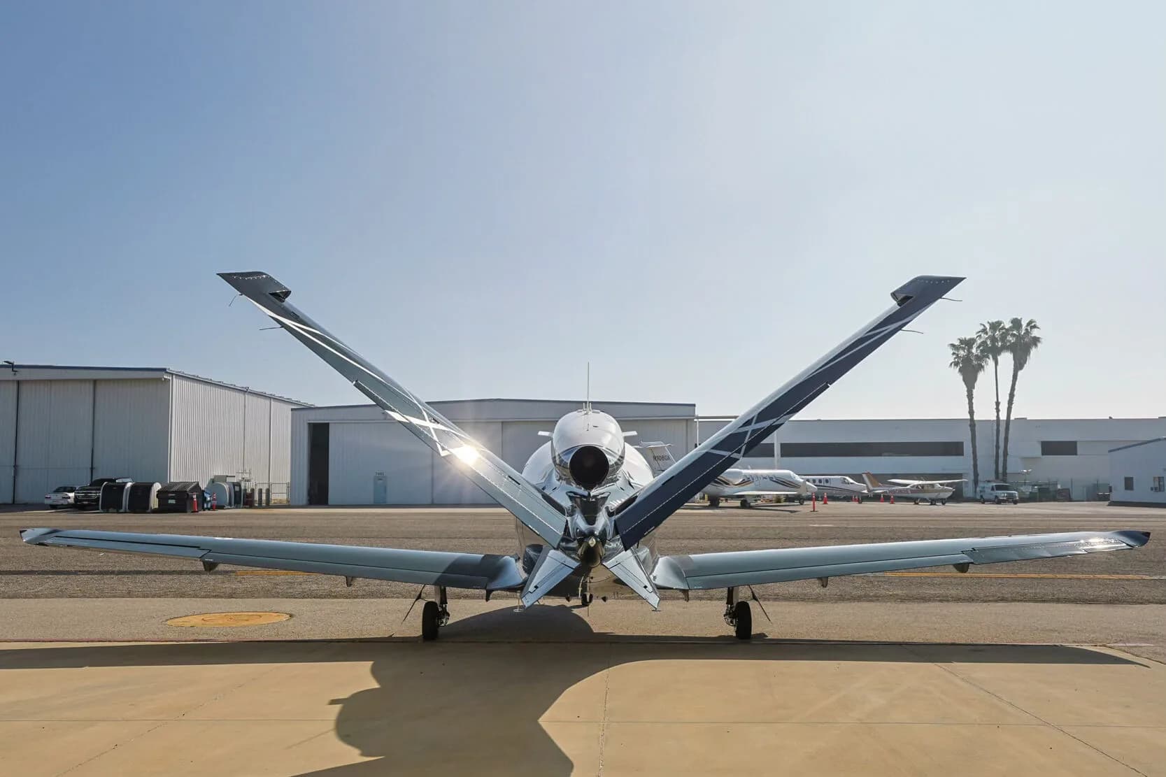 Rear view of a Cirrus Vision Jet with its distinctive V-tail, parked on an tarmac with palm trees in the background.