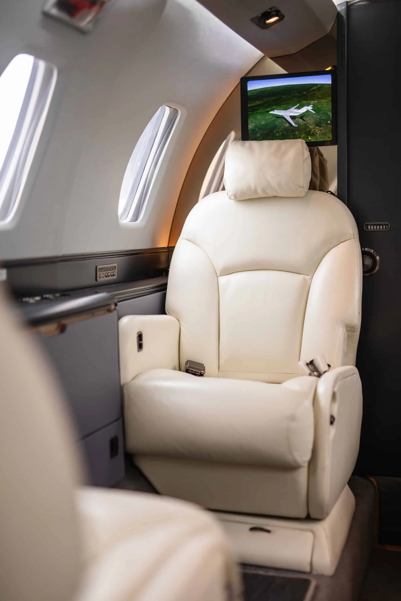 A single light-colored leather seat in a private jet cabin with a personal entertainment screen showing an aircraft in flight.