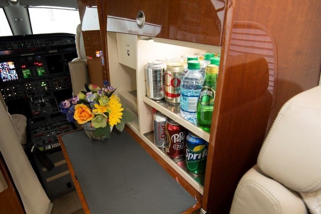 A private jet's refreshment center stocked with various beverages, with the cockpit visible in the background.