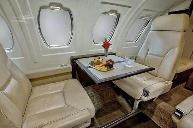 A close-up view of the private jet's interior cabin shows light leather seats and a table with a meal and a red rose.
