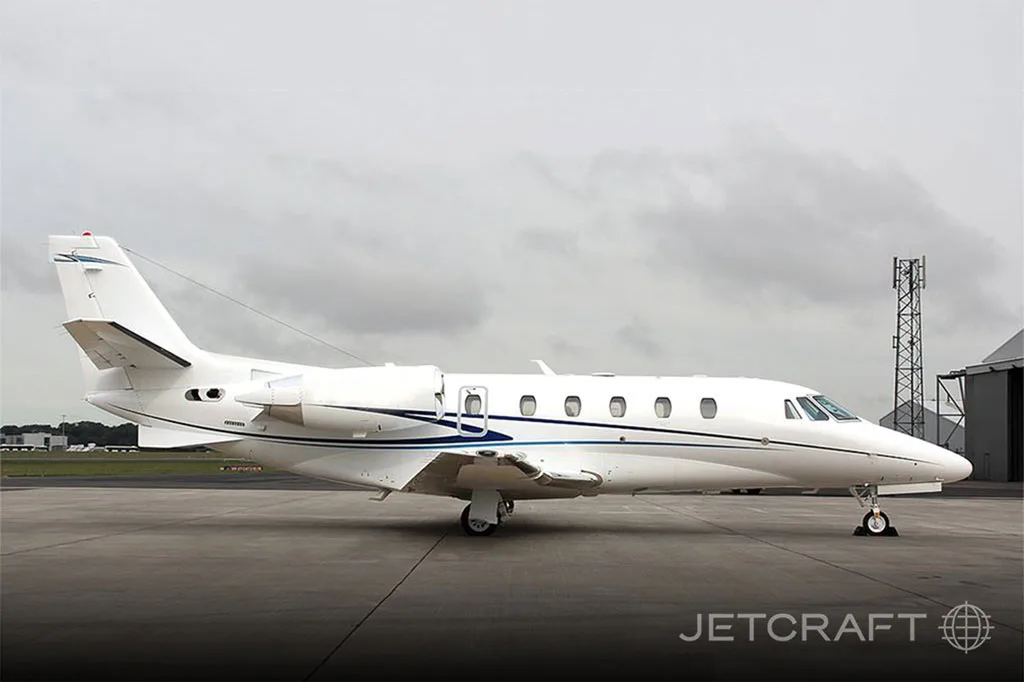 A white business jet with blue accent stripes is parked on an asphalt apron under a cloudy sky.