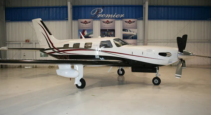 A white and black Piper Meridian aircraft with red accents, registration N3CV, parked in a hangar with "Premier" and "Mooney" banners.