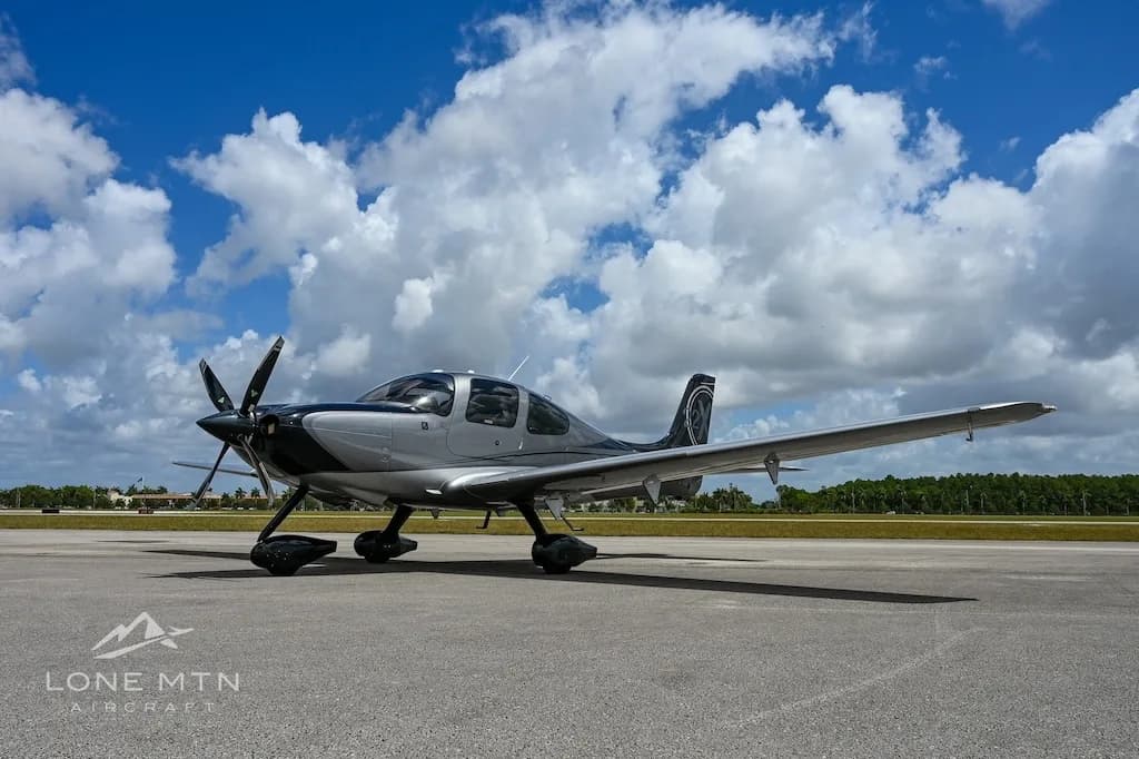 A silver and black Cirrus SR22 aircraft parked on an airfield under a partly cloudy sky, viewed from the front-left.