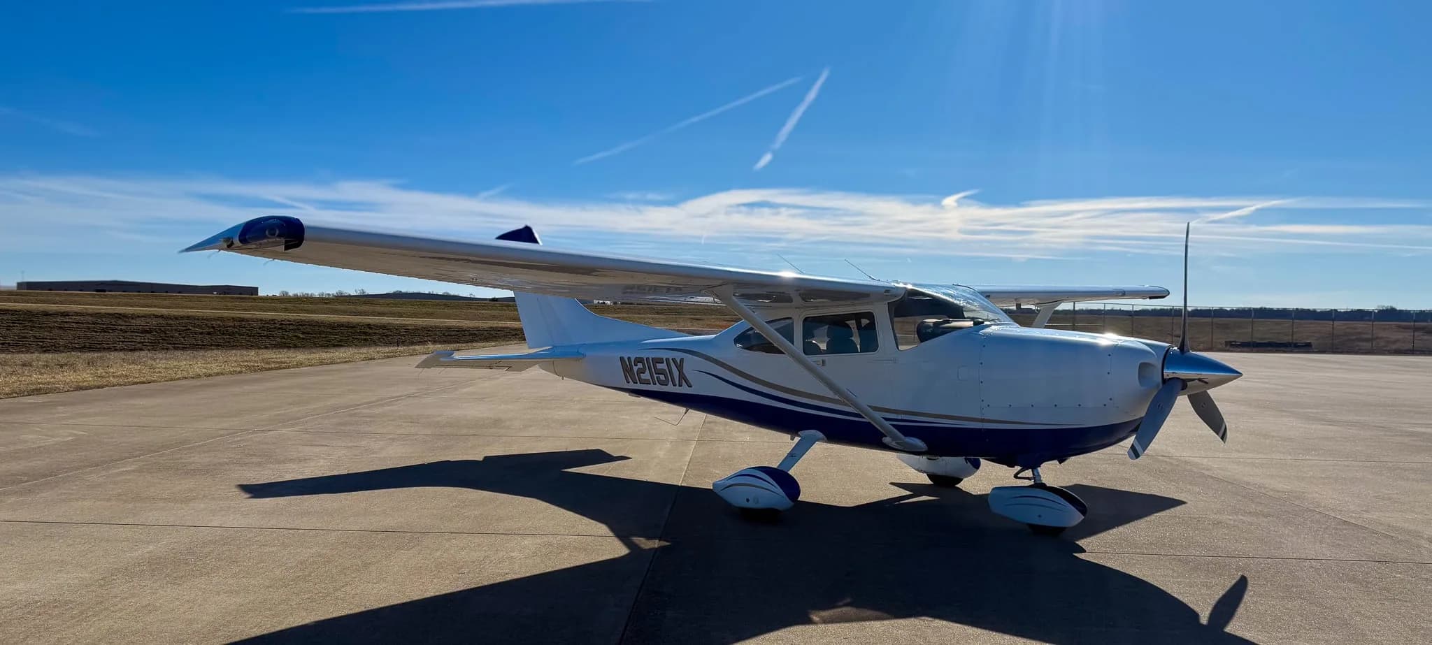 A white and blue Cessna 182 Skylane (N2151X) parked on a tarmac under a clear blue sky, viewed from the front-right.