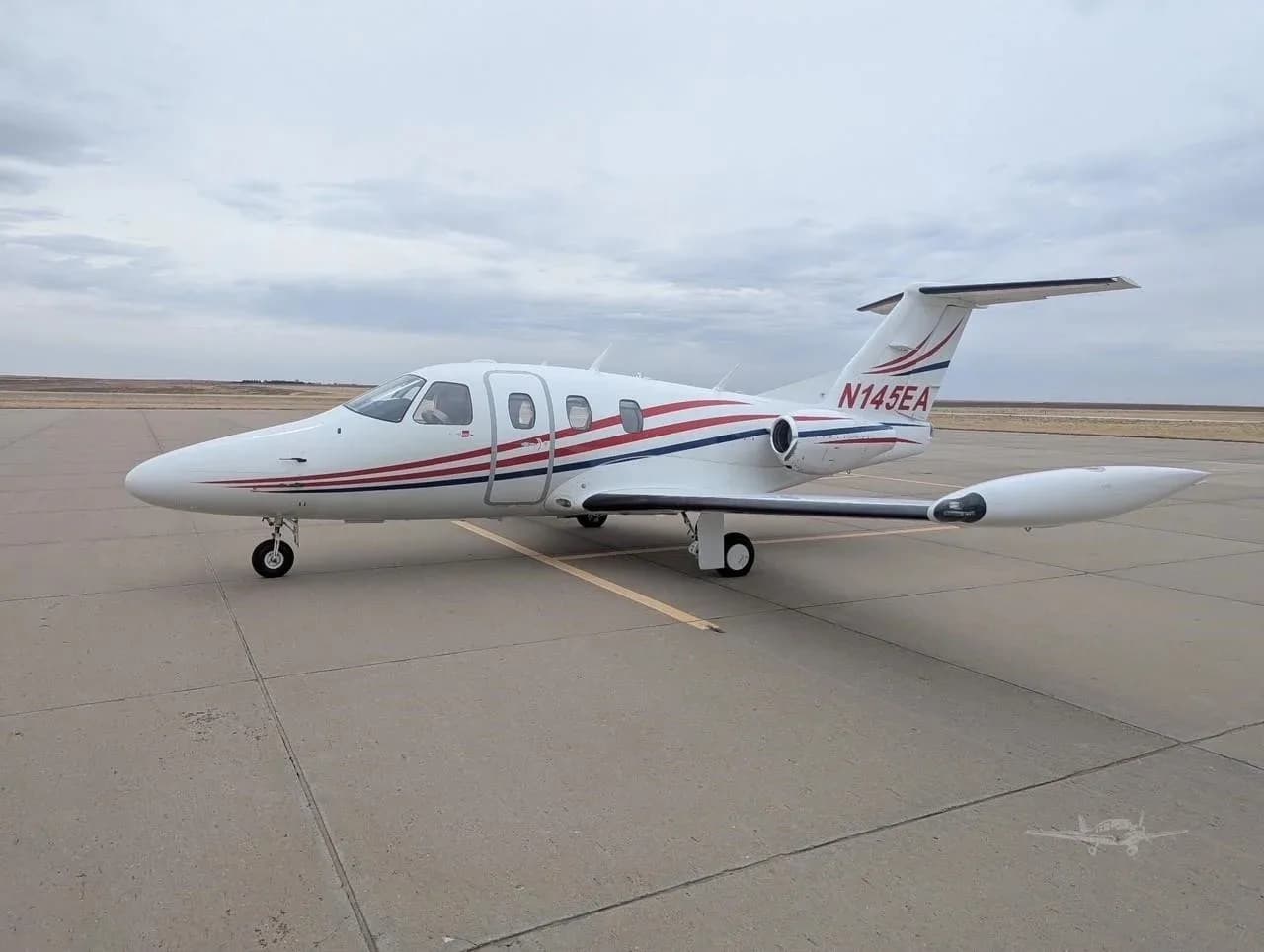 A white Eclipse jet with red and blue stripes, registration N145EA, parked on an airfield under a cloudy sky.