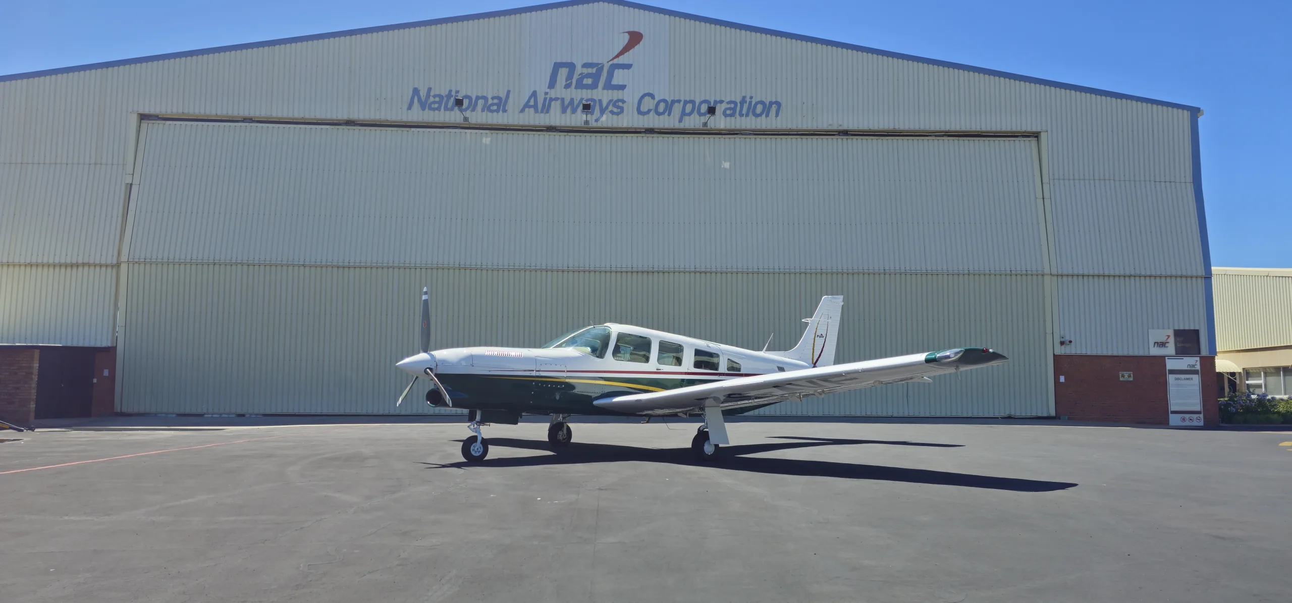 A white and dark green Piper single-engine aircraft parked on an apron in front of a National Airways Corporation hangar.